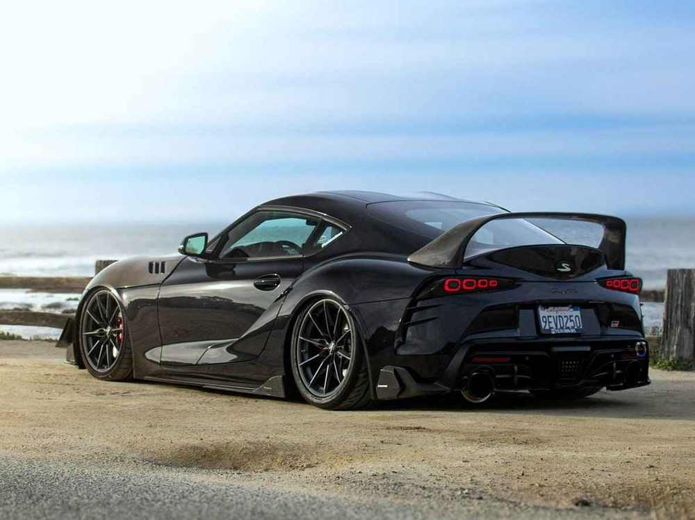A black Toyota Supra with ARMASPEED carbon fiber kit parked at a coastal road, rear three-quarter view.
