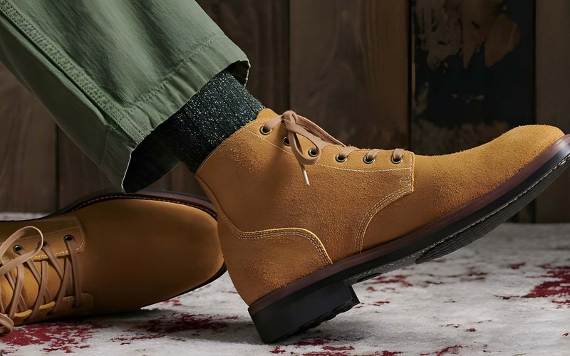 A tan suede chukka boot on green chinos against a dark wood-plank backdrop — SuperDry AW20 chukka line.
