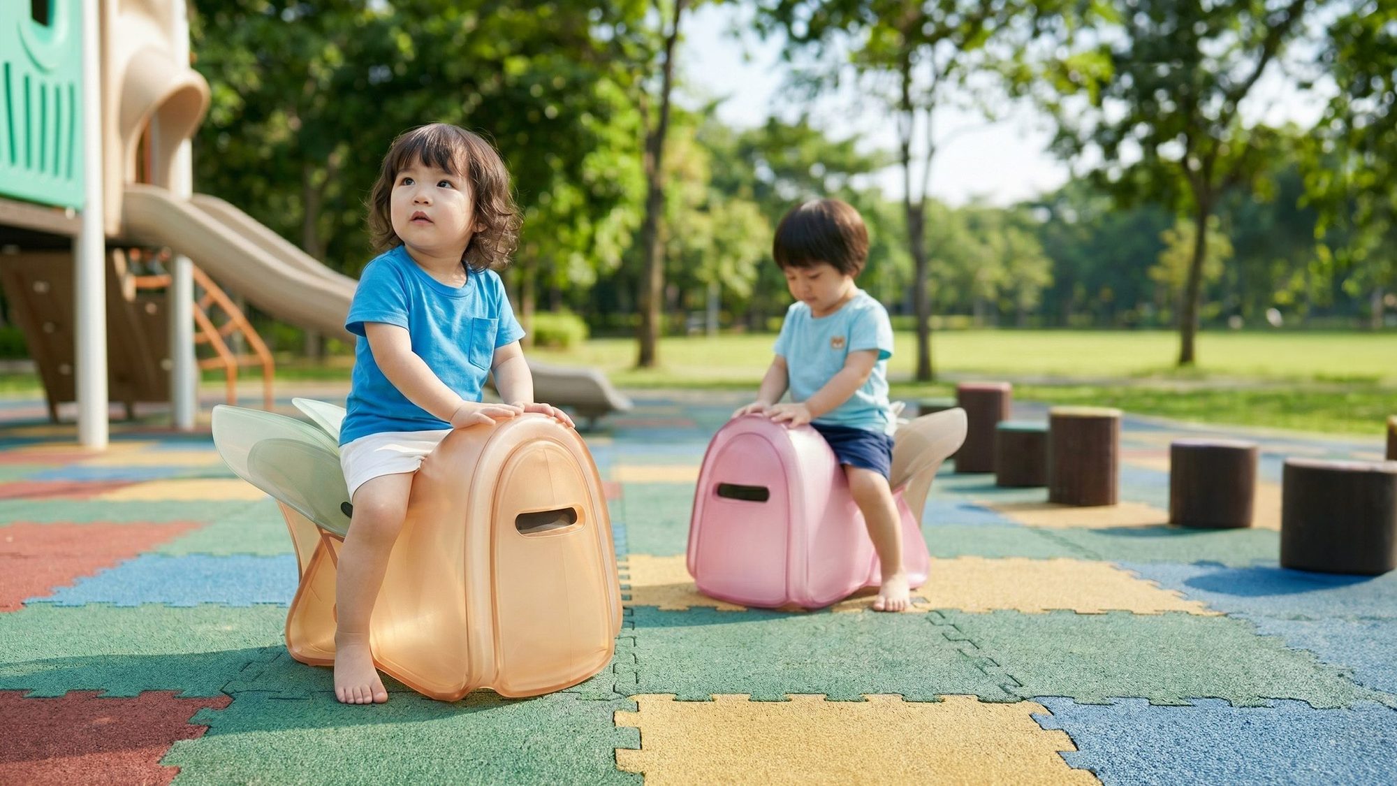 Two Taiwanese children sitting on cicada-shaped chair-cajóns on a rubber-tile playground floor, with a playground slide and banyan trees in the background.